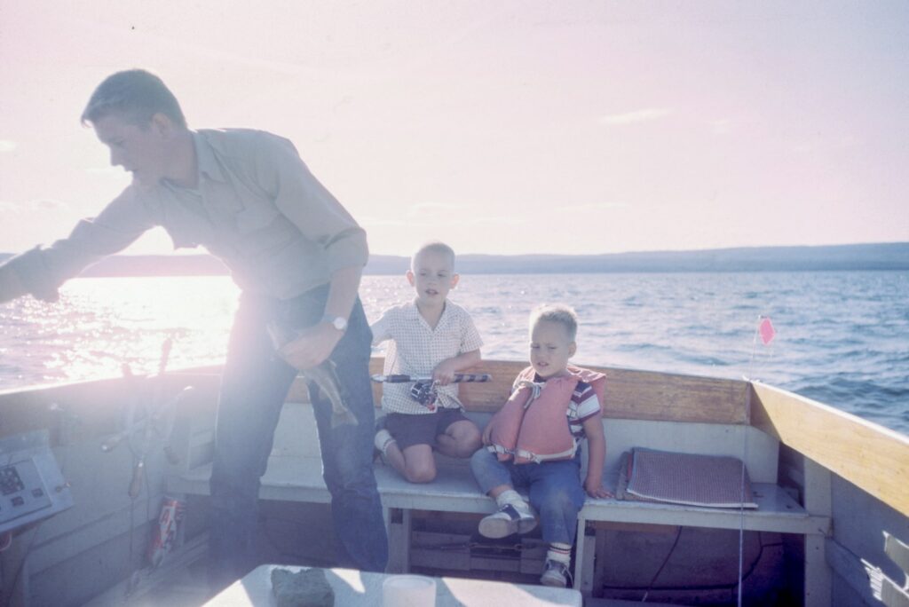 man holding gray fish standing beside two boys sitting on boat seats