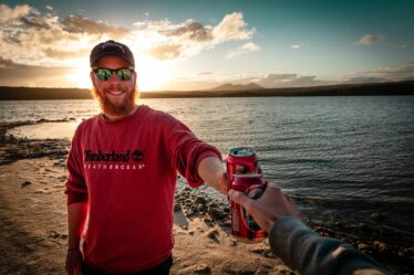 man in red sweater toasting red beverage can near body of water during daytime